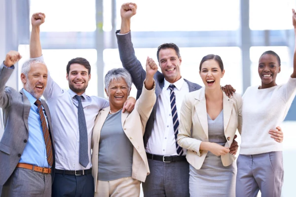 Diverse group of business professionals celebrating success with raised arms and smiles in a bright office setting.