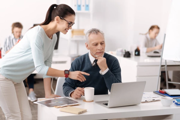 Young woman assisting senior man with laptop at office desk. Other coworkers are visible in the background.