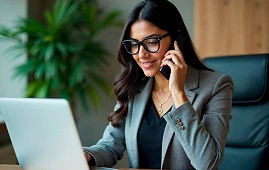 Businesswoman with glasses using laptop and talking on phone in office setting.