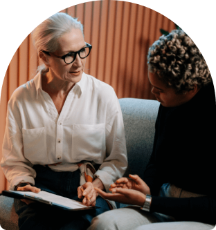 Mature woman in glasses listens intently to a younger person. They are seated and discussing a document on a clipboard.