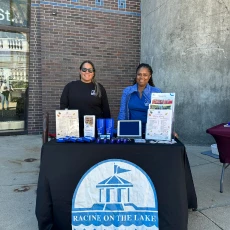 Two women staffing a Racine On The Lake booth at an outdoor event. Table with brochures, tablet, and branded items.