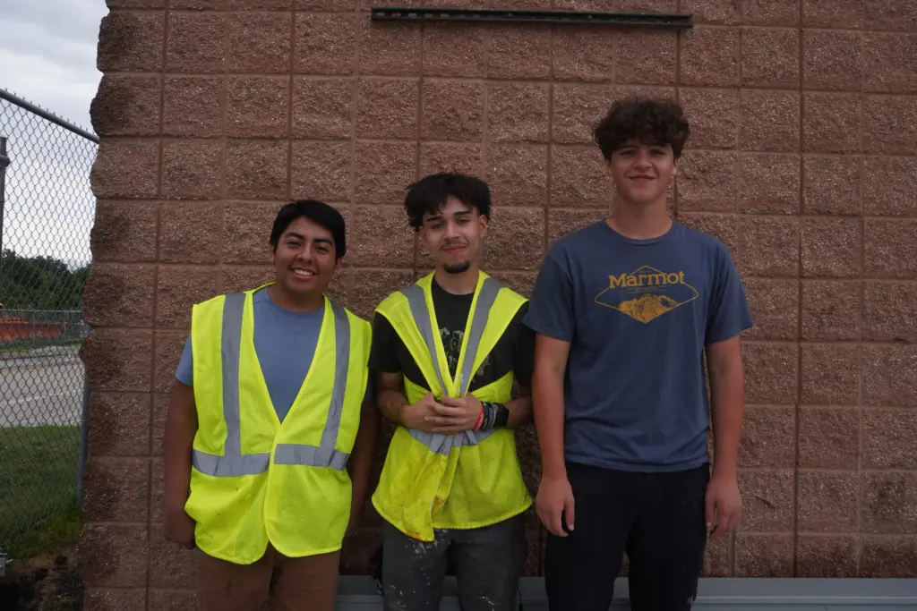 Three young men stand in front of a brick building, two wearing bright yellow safety vests.