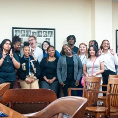 Group of diverse students posing for a photo in a room with wooden chairs and framed pictures on the wall.