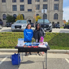 Two women stand behind a table with items for distribution outside a building, likely a community event.
