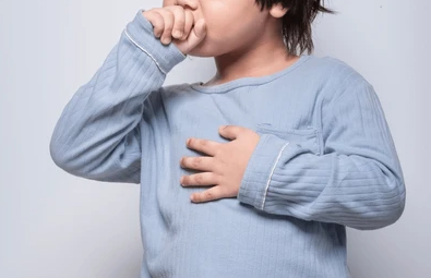 A young child wearing a light blue sweater who appears to be coughing while covering their mouth.